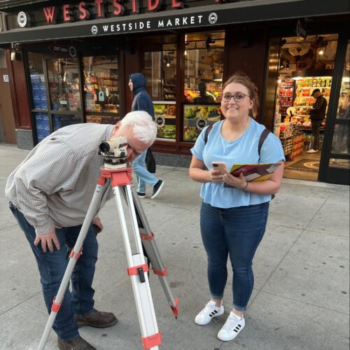 A student and instructor conduct a surveying exercise on a city sidewalk outside Westside Market. The instructor looks through a leveling instrument mounted on a tripod, while the student stands nearby holding a phone and clipboard, smiling at the camera.
