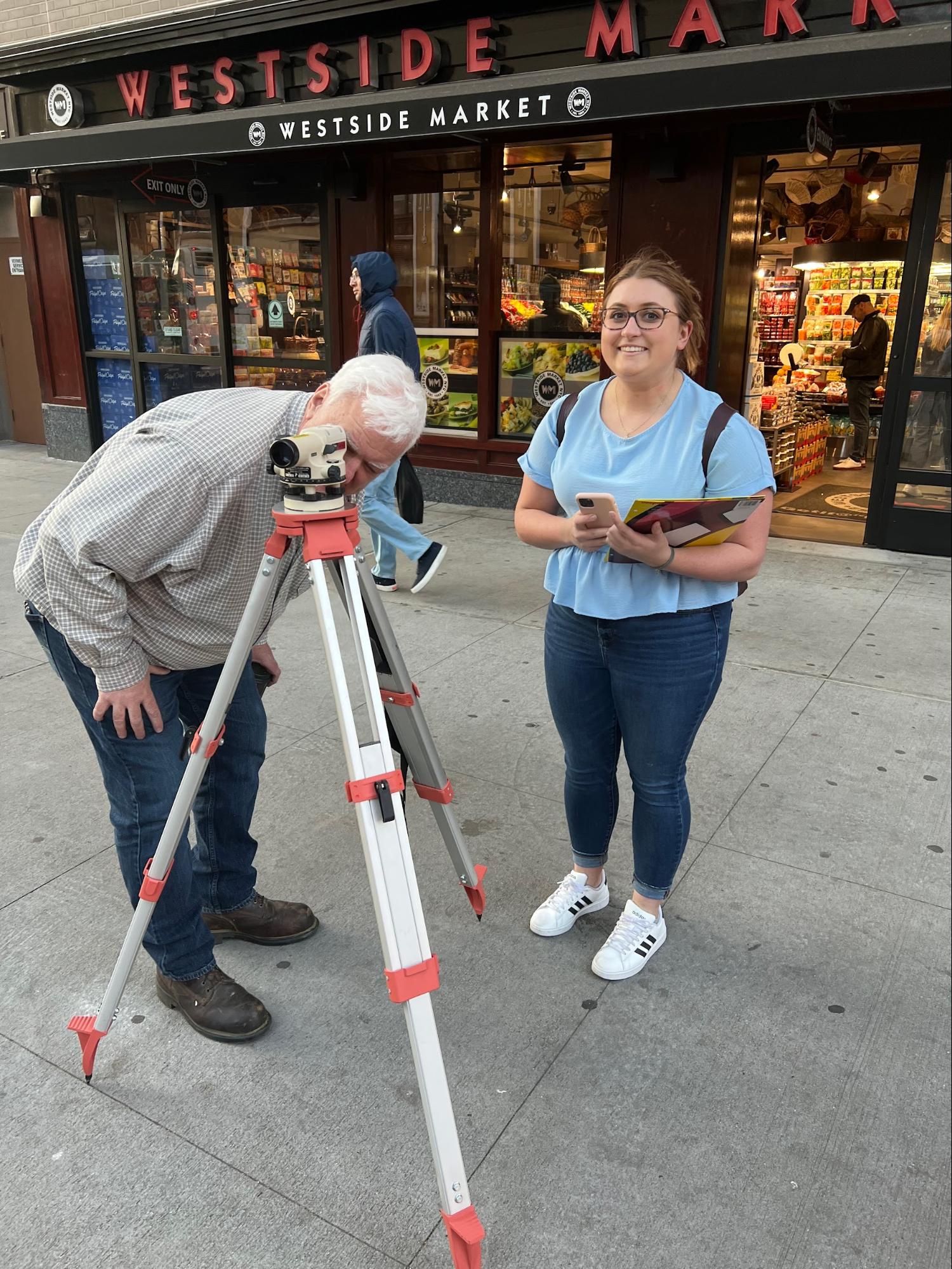 A student and instructor conduct a surveying exercise on a city sidewalk outside Westside Market. The instructor looks through a leveling instrument mounted on a tripod, while the student stands nearby holding a phone and clipboard, smiling at the camera.