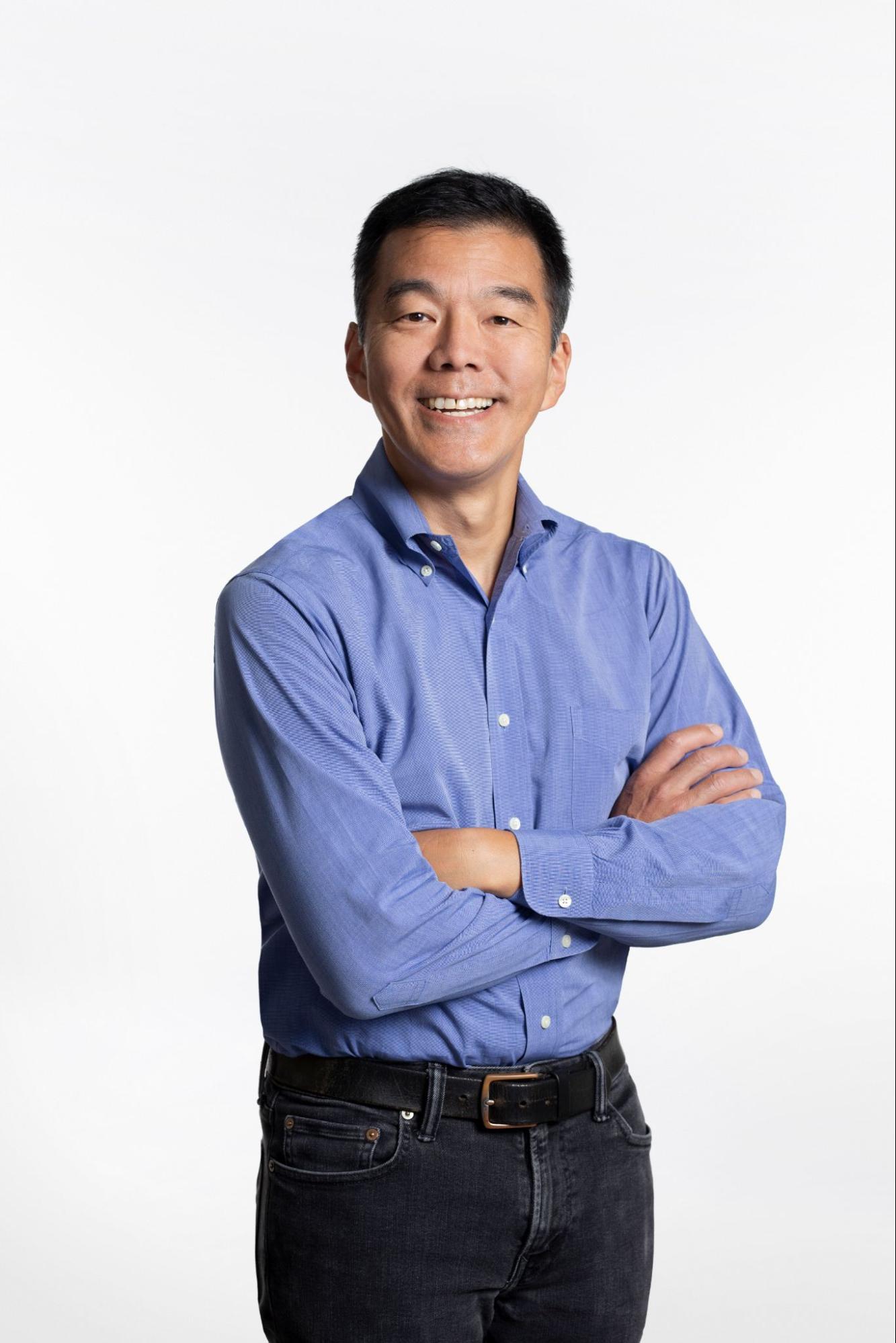 Portrait of a man smiling with arms crossed, wearing a blue button-down shirt and black jeans, standing against a plain white background.