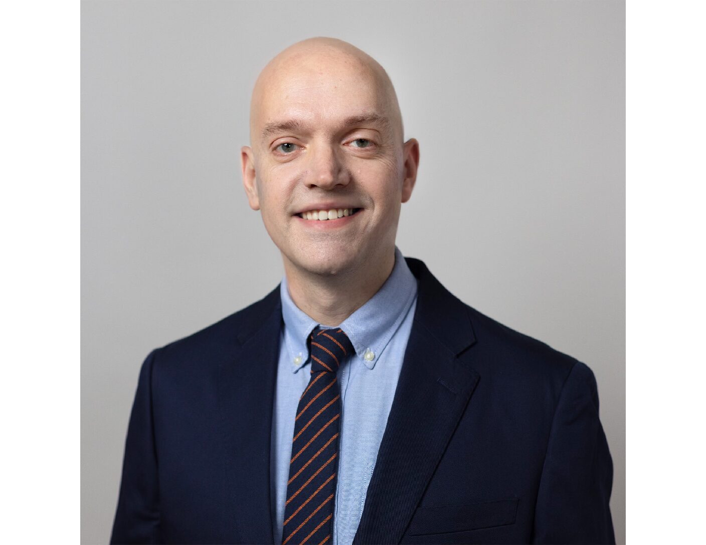 Professional headshot of a man wearing a navy blue suit, light blue shirt, and striped tie, smiling against a neutral gray background.