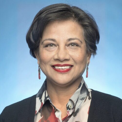 Headshot of a woman smiling warmly against a blue background, wearing a patterned blouse, black cardigan, and red earrings.