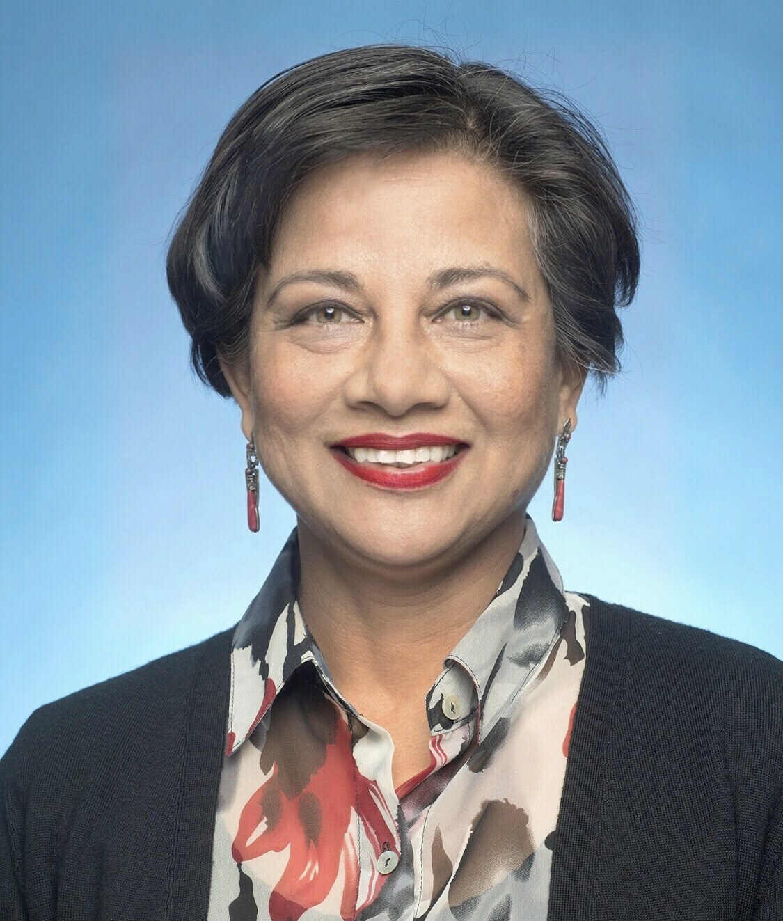 Headshot of a woman smiling warmly against a blue background, wearing a patterned blouse, black cardigan, and red earrings.