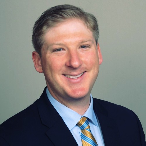Professional headshot of a man with short light brown hair, wearing a dark suit, light blue shirt, and yellow-striped tie, smiling against a neutral background.