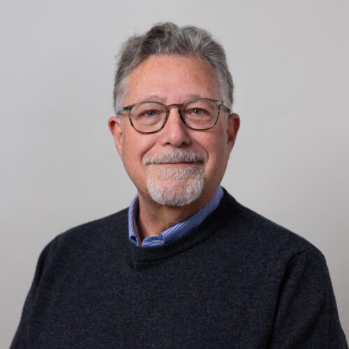 Professional portrait of an older man with gray hair, glasses, and a goatee, wearing a dark sweater over a collared shirt, smiling softly against a neutral light gray background.