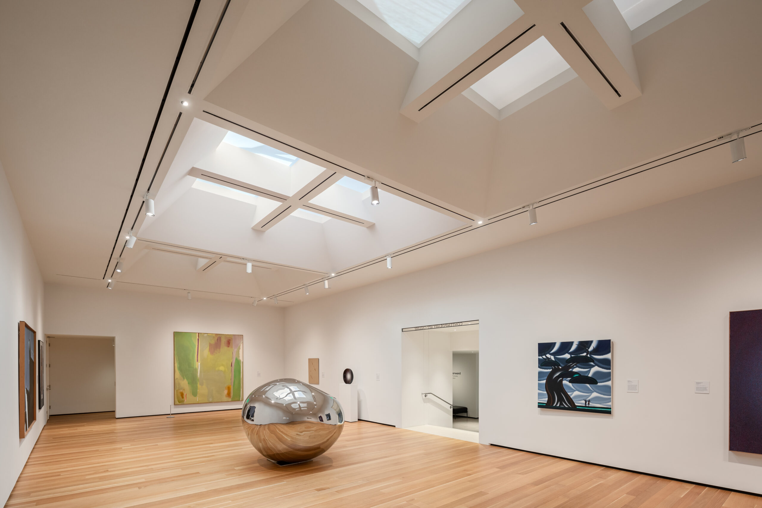 A white gallery space with windows in the ceiling, paintings on the walls, and a amorphous globule made of reflective metal.