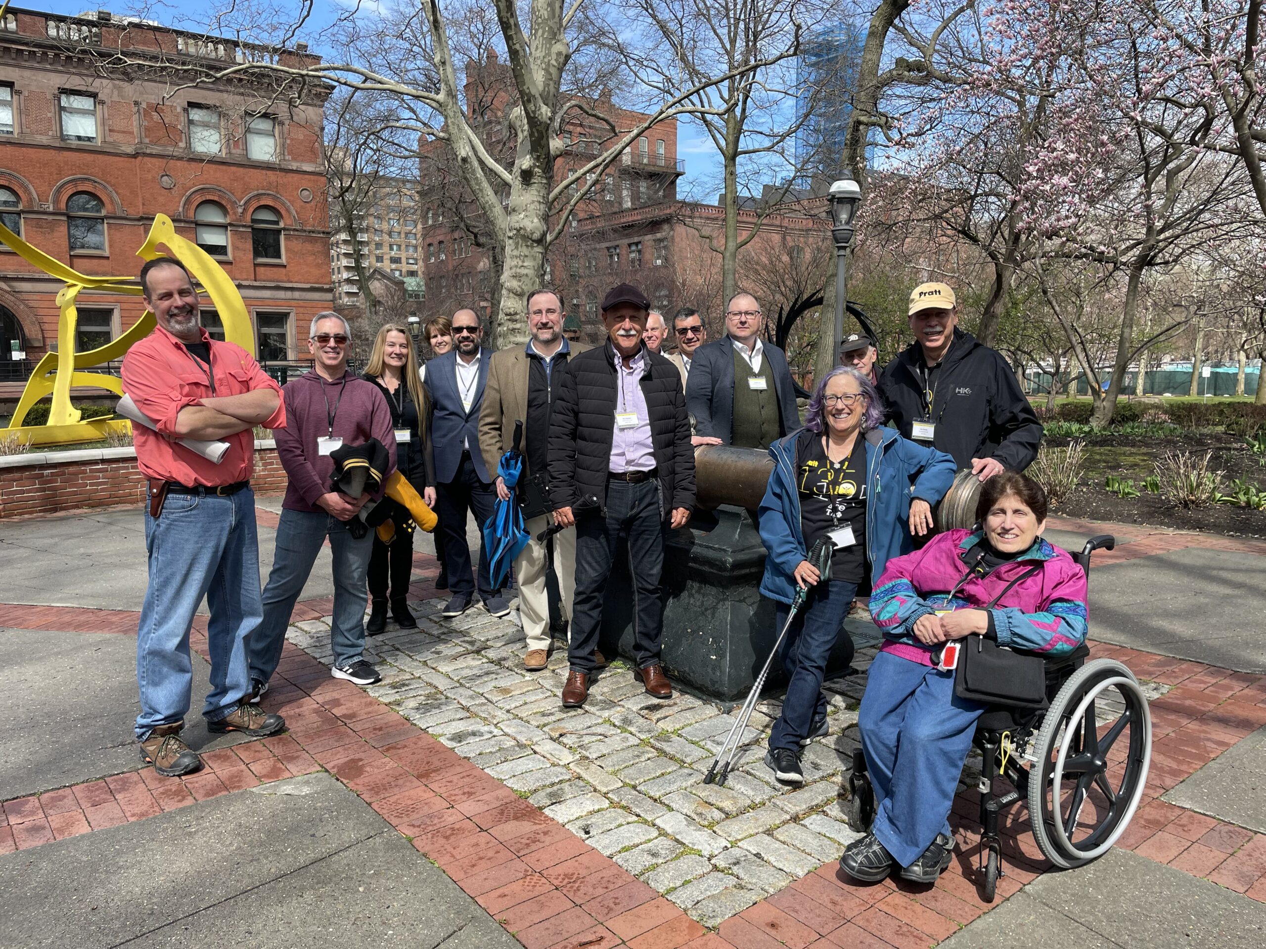 A photograph of fourteen people standing outside and posing for a group portrait. They are standing on a paved surface in Pratt’s campus, with trees, sculptures, and buildings in the background.