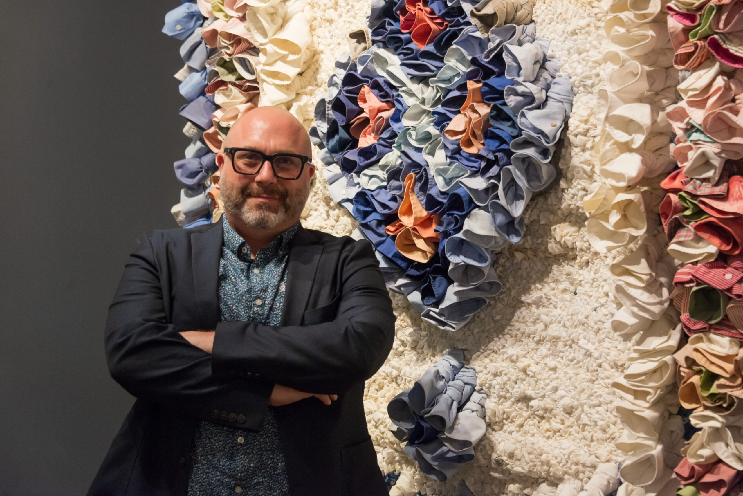 A photograph of a man crossing his arms in front of fabric-based fine art.