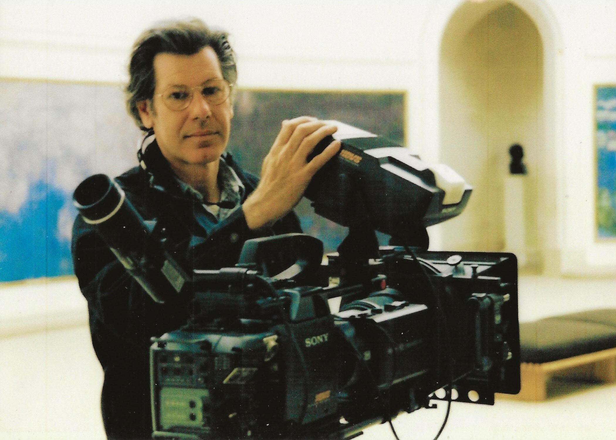 A man in sunglasses holding a large film camera on a tripod in a gallery space.