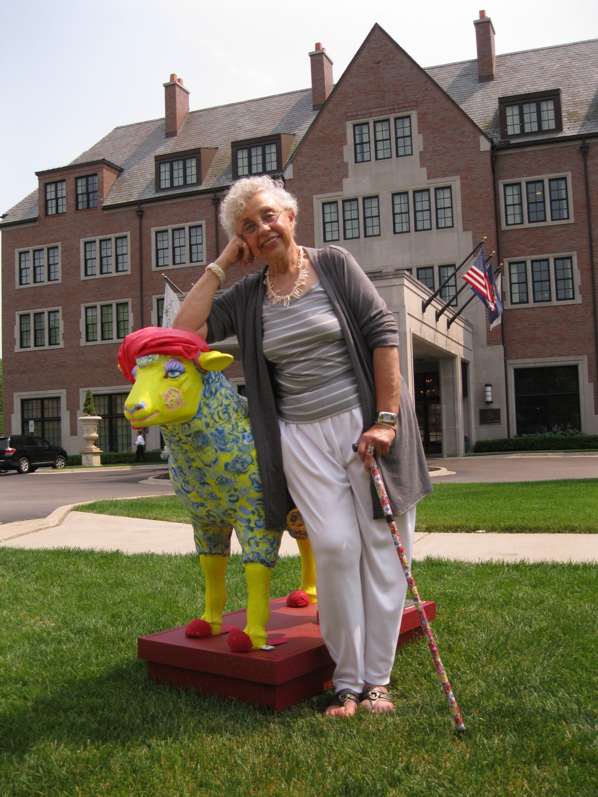 A woman with a cane leaning on a colorful statue of a sheep on a plot of grass in front of a building.