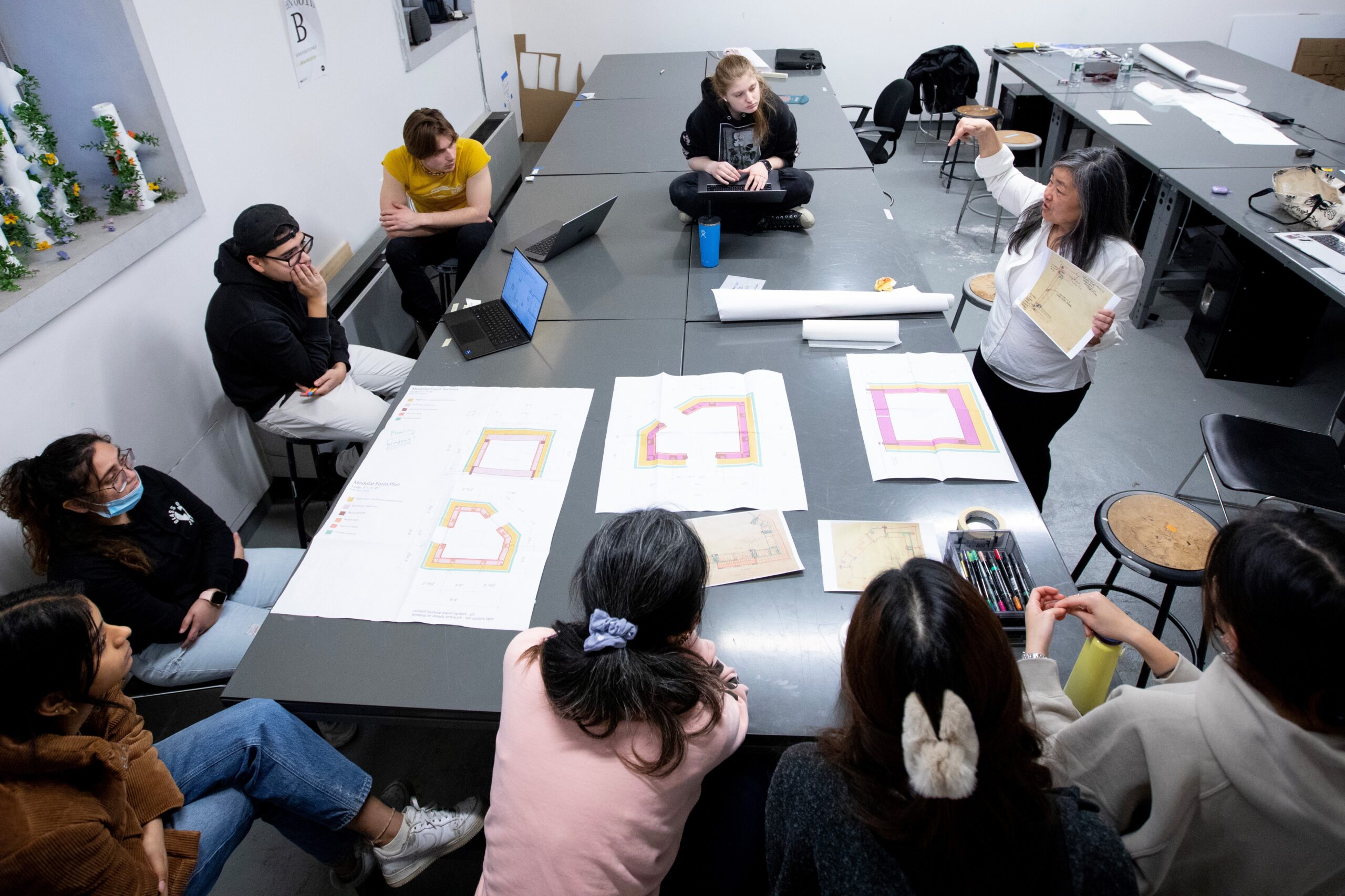 Professor In Cho presents at a large table where eight students in a circle look at architectural drawings