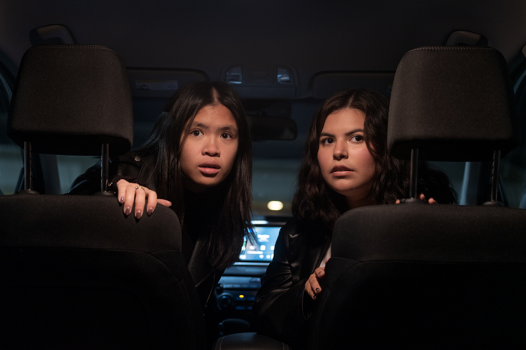 Photo of two girls taken from the second row of a car. The girls are sitting in the front seats but are turned backwards towards the camera. They are looking past the camera in disbelief.