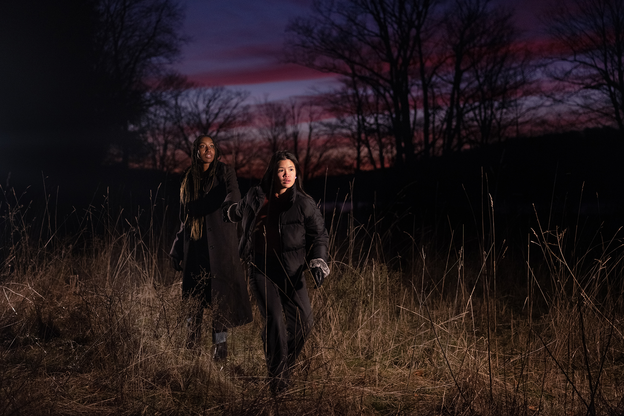 Two girls walk amongst tall grass. One is leading the other by the hand as they both look up to the sky in awe.