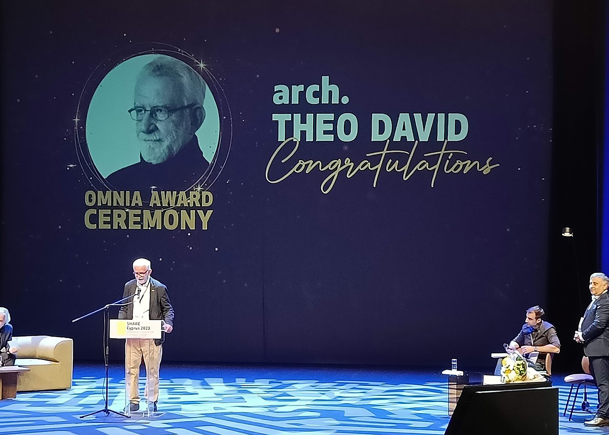A man on a blue stage behind a podium and in front of a screen which reads “Omnia Award Ceremony: Architect Theo David: Congratulations!” There are three people on stage who are watching, sitting on or standing beside chairs and couches.