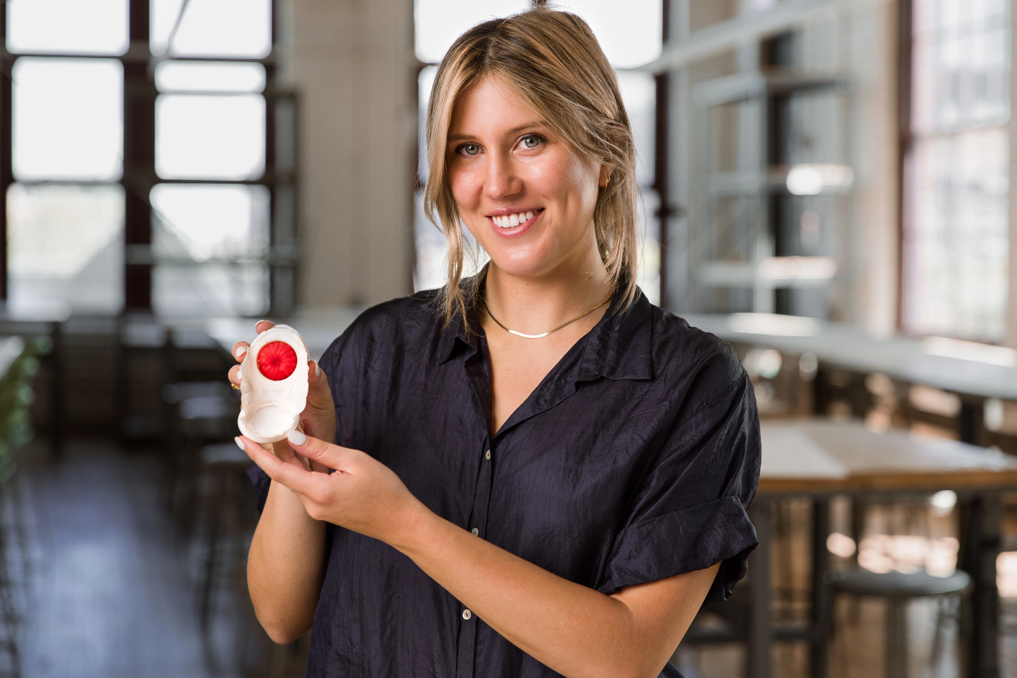 A woman smiles and poses for the camera. She is holding a small white and red device in her hands displaying it for the viewer.