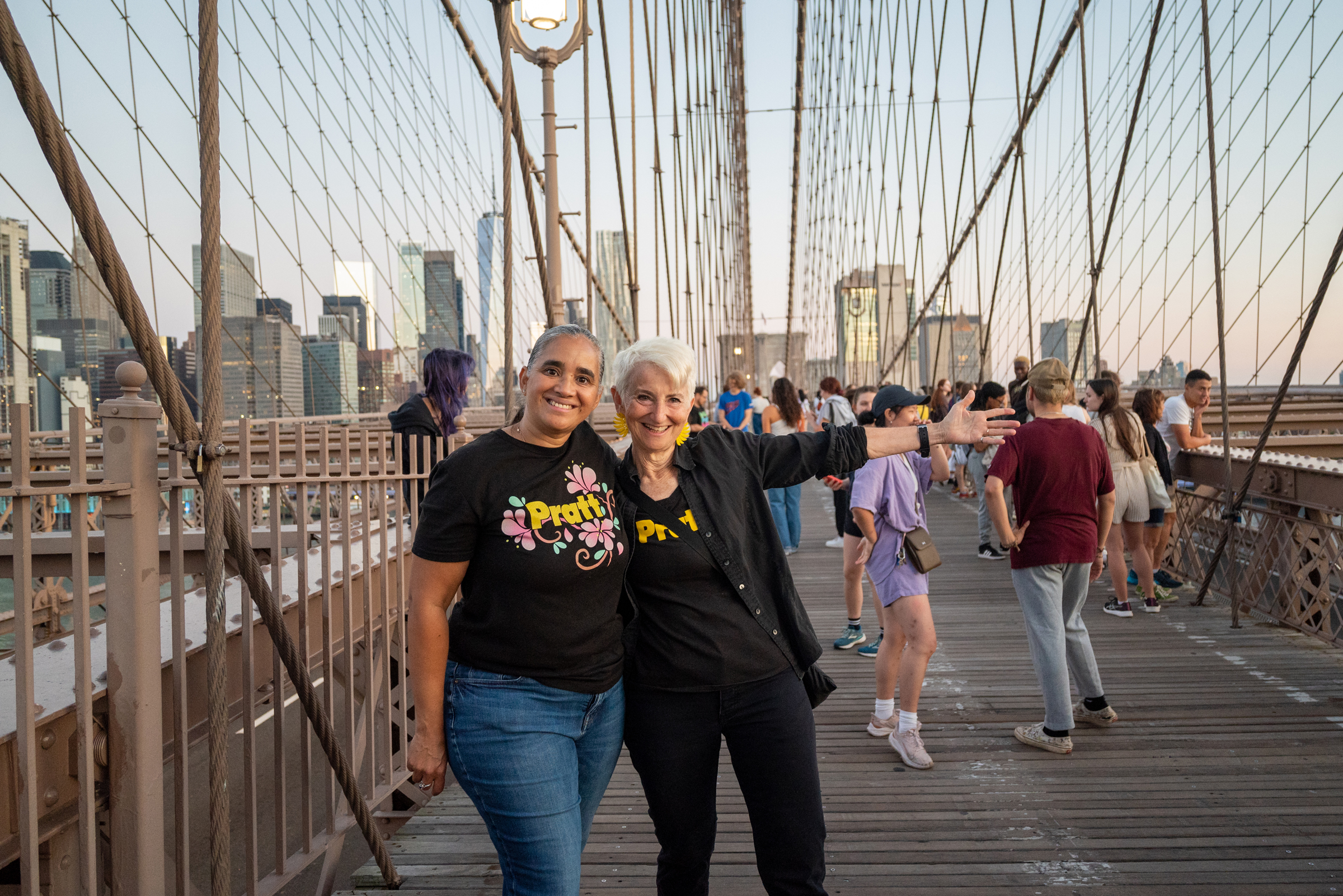 President Bronet and another staff member are on the Brooklyn Bridge looking at the camera. They are wearing t-shirts with the Pratt logo. The New York City skyline is behind them and students are looking out over the side of the bridge.