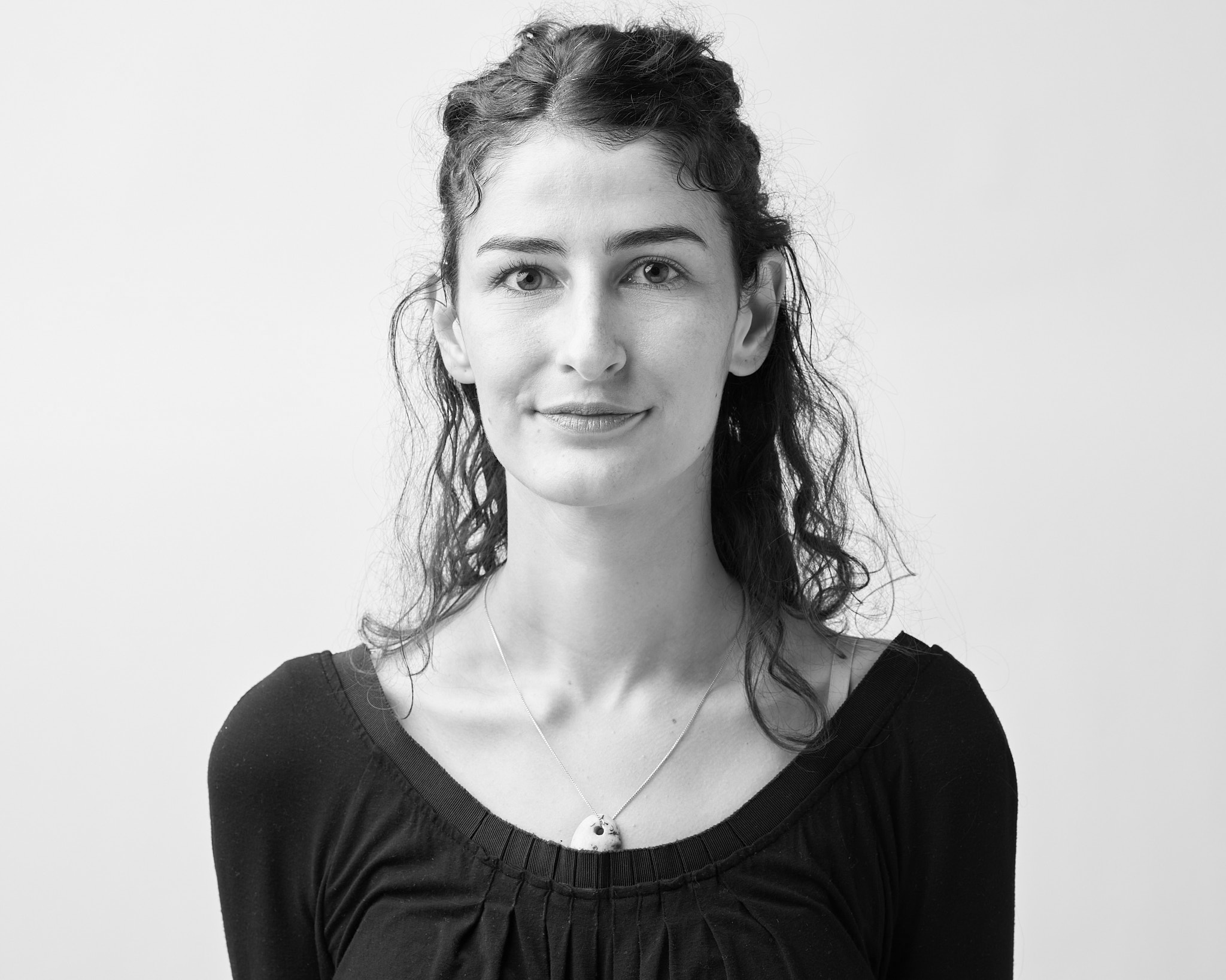 Black-and-white studio portrait of an adult person with curly shoulder-length hair, wearing a light blouse and facing slightly toward the camera against a plain light background.