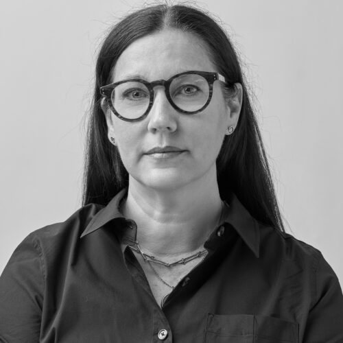 Black-and-white studio portrait of a woman with long straight hair wearing round glasses and a dark button-up shirt, looking directly at the camera against a plain light background.