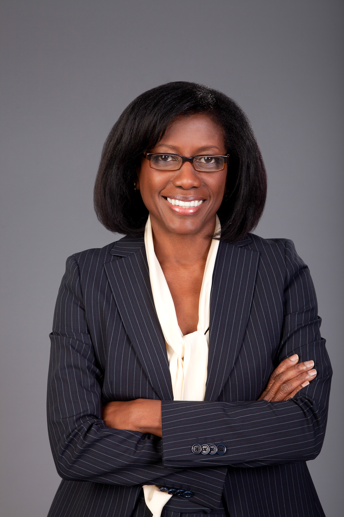 Woman in blazer and glasses smiles with arms crossed in a professional portrait