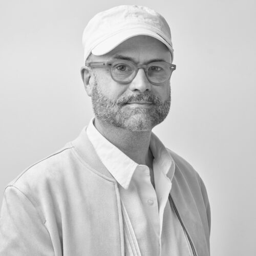 Black-and-white studio portrait of a bearded man wearing glasses, a light-colored baseball cap, and a casual jacket over a button-up shirt, facing the camera against a plain light background.