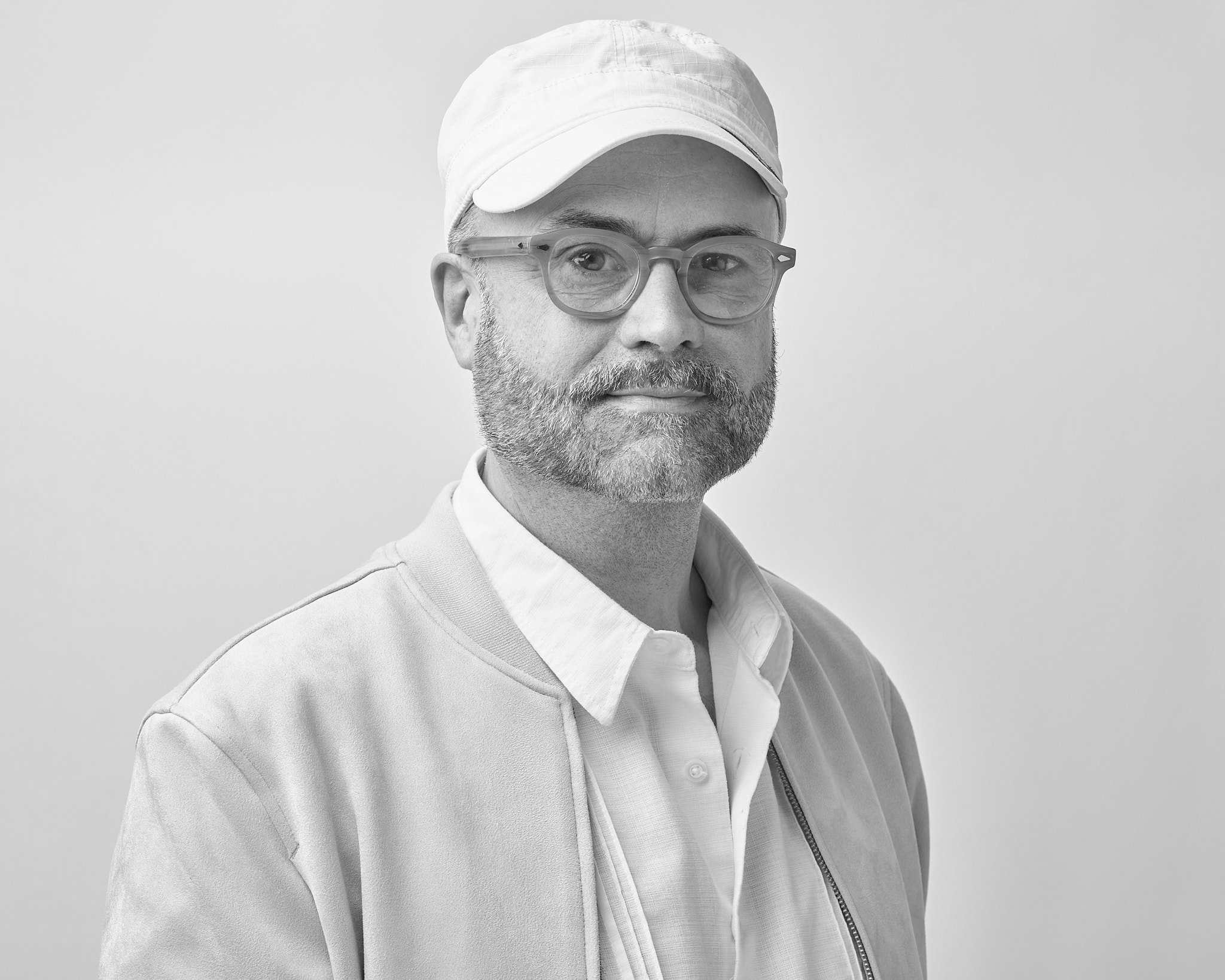 Black-and-white studio portrait of a bearded man wearing glasses, a light-colored baseball cap, and a casual jacket over a button-up shirt, facing the camera against a plain light background.