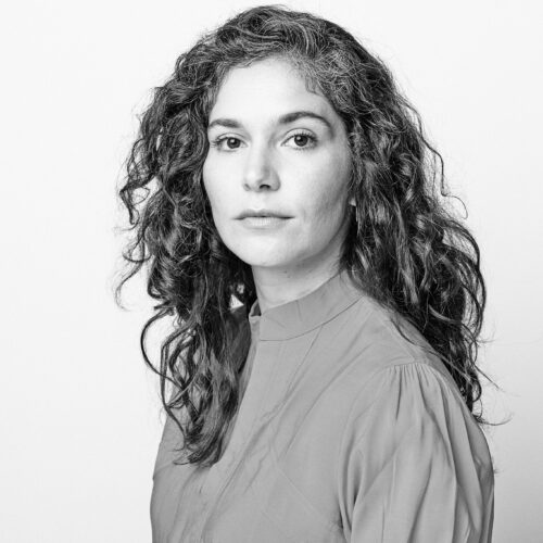 Black-and-white studio portrait of an adult person with long hair pulled back over one shoulder, wearing a dark top and looking directly at the camera against a plain background.