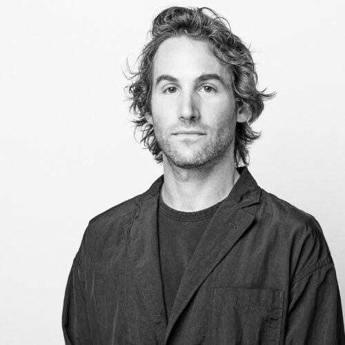 Black-and-white studio portrait of an adult person with short hair and a trimmed beard, wearing glasses and a dark jacket, facing the camera against a plain light background.