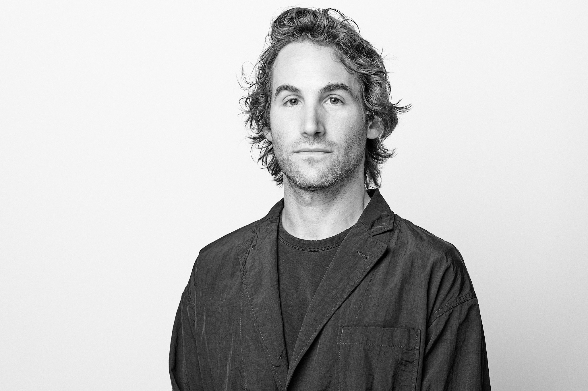 Black-and-white studio portrait of an adult person with short hair and a trimmed beard, wearing glasses and a dark jacket, facing the camera against a plain light background.