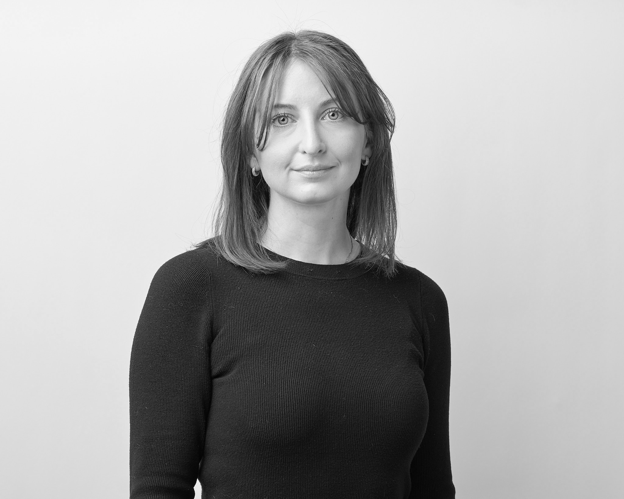 Black-and-white studio portrait of an adult person with shoulder-length hair and bangs, wearing a dark long-sleeve top and looking calmly at the camera against a plain background.