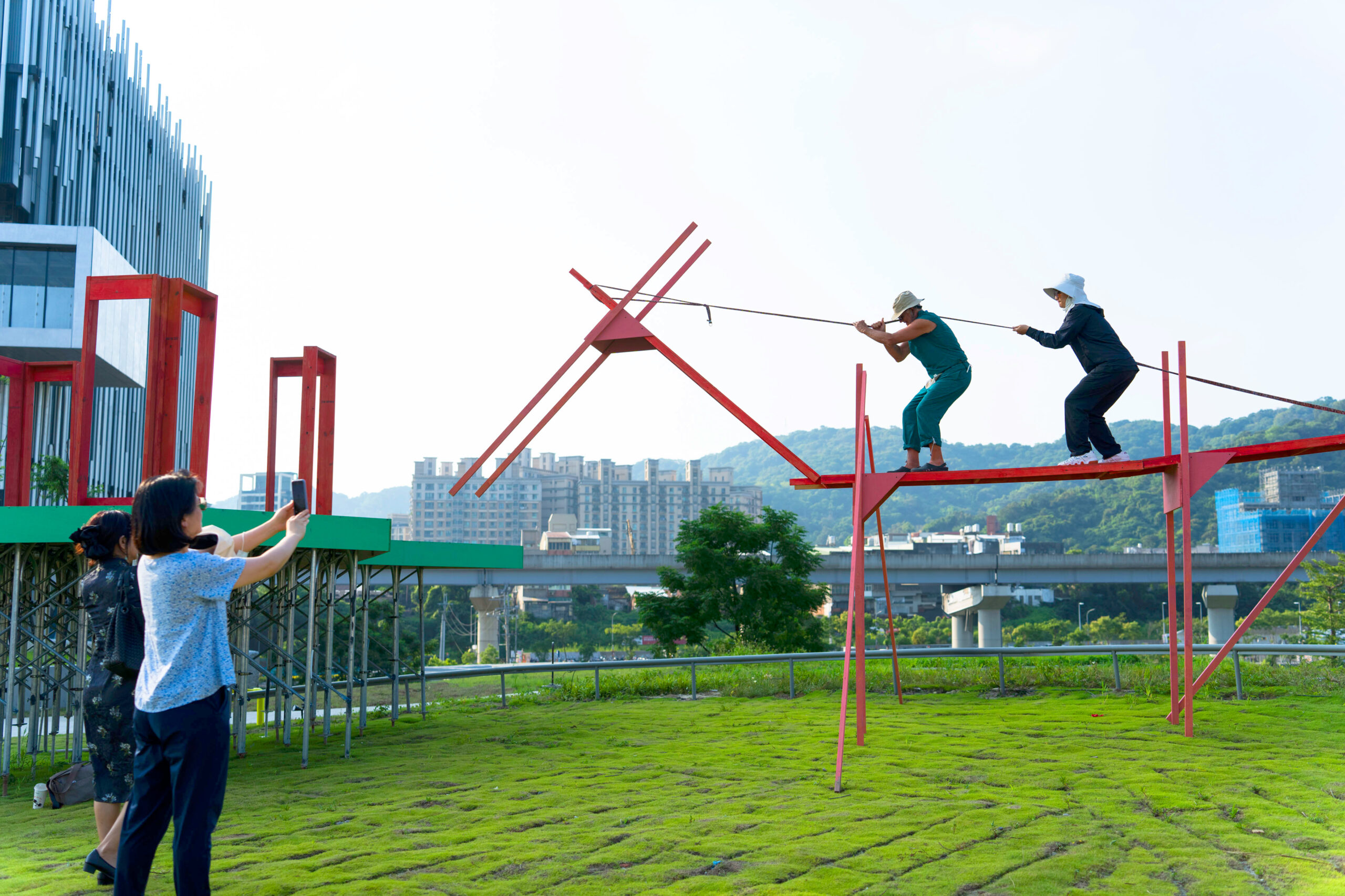 Two people lower a plank on long legs toward the ground in front of platform with three rectangular structures