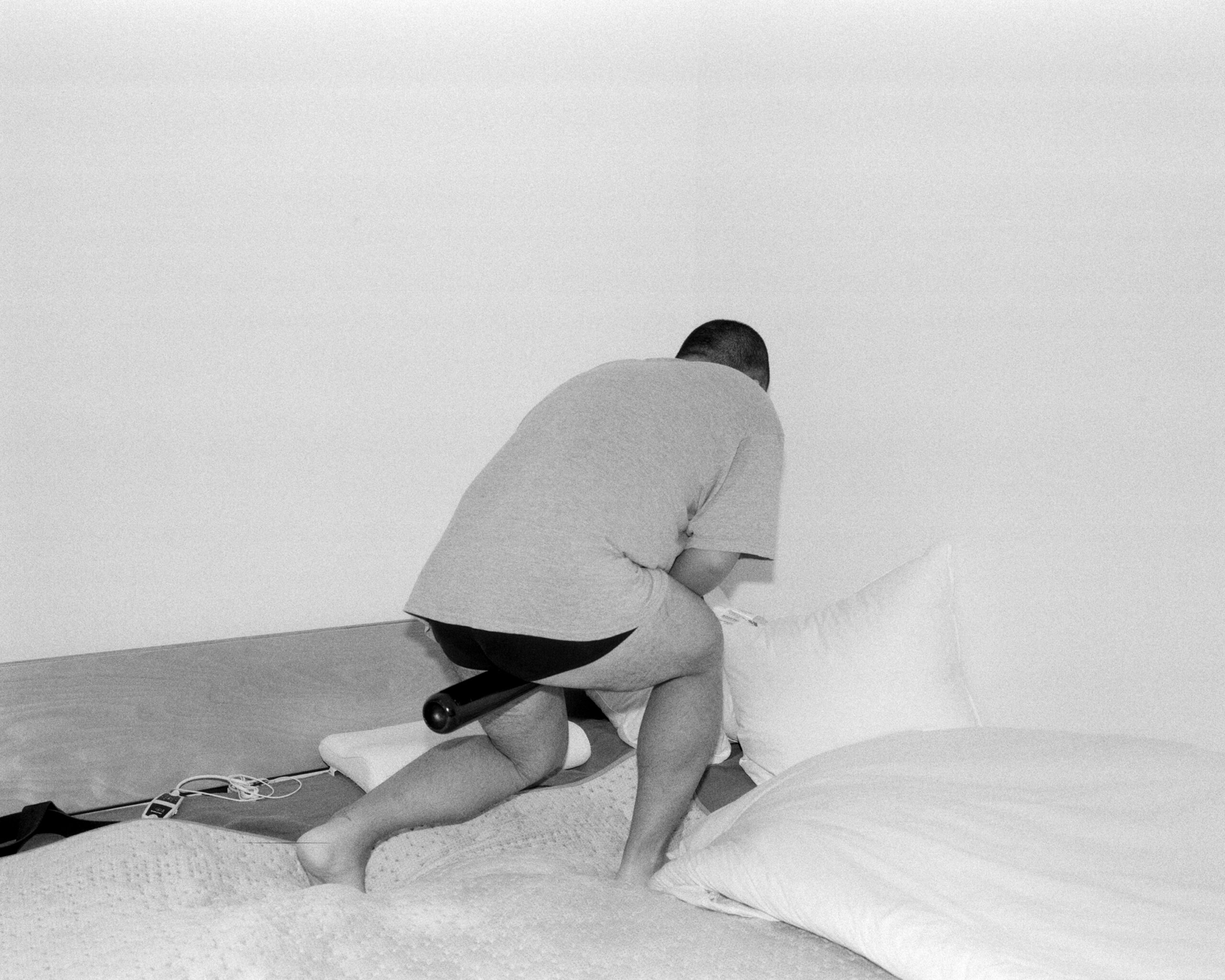 Black-and-white photograph of a person with short hair in black briefs and T-shirt standing back to the camera on a bed with a black cylinder protruding from between their legs