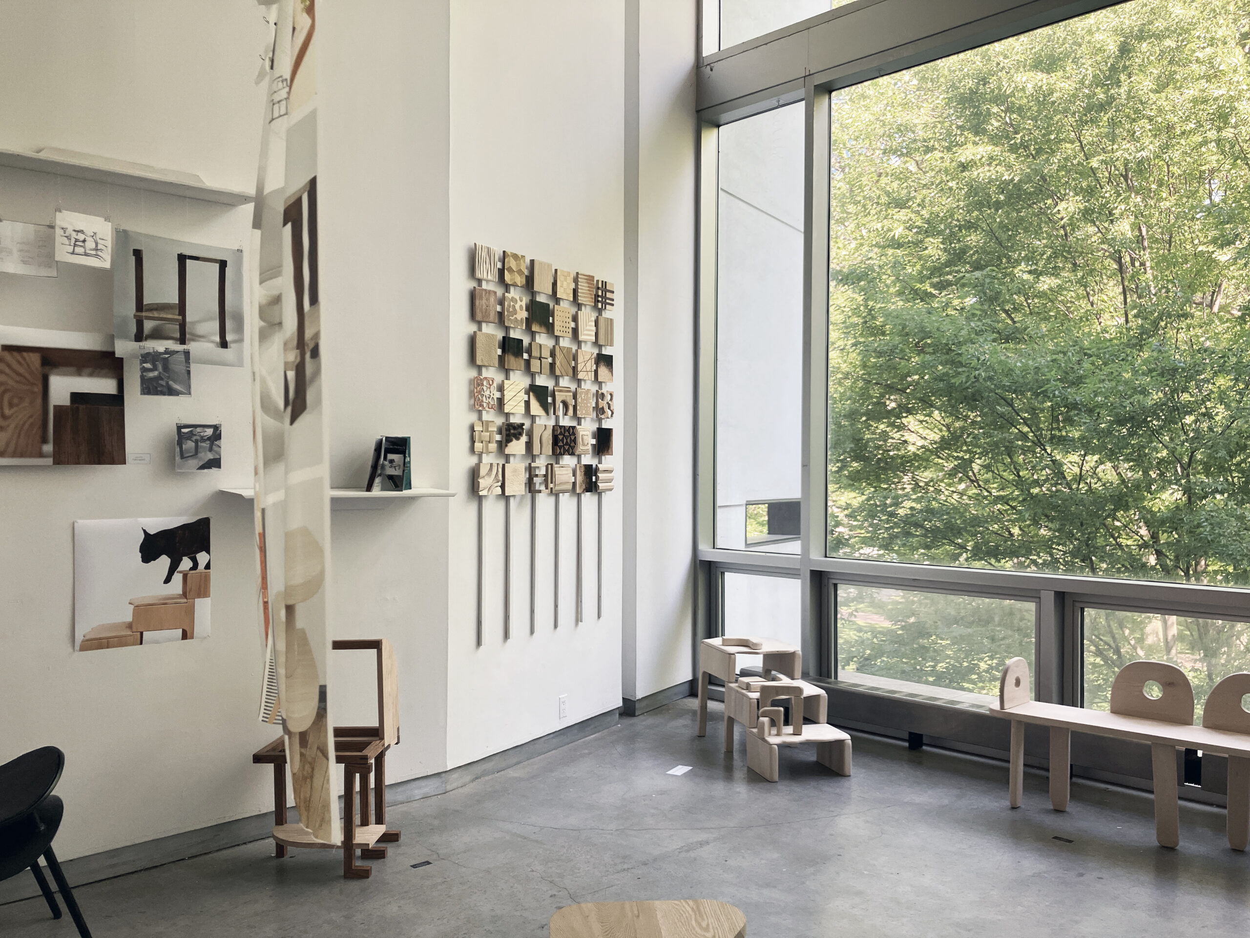 Corner of gallery with wall-size window flooding natural light onto installation of furniture, wall installation of wood blocks, and other design-process documentation