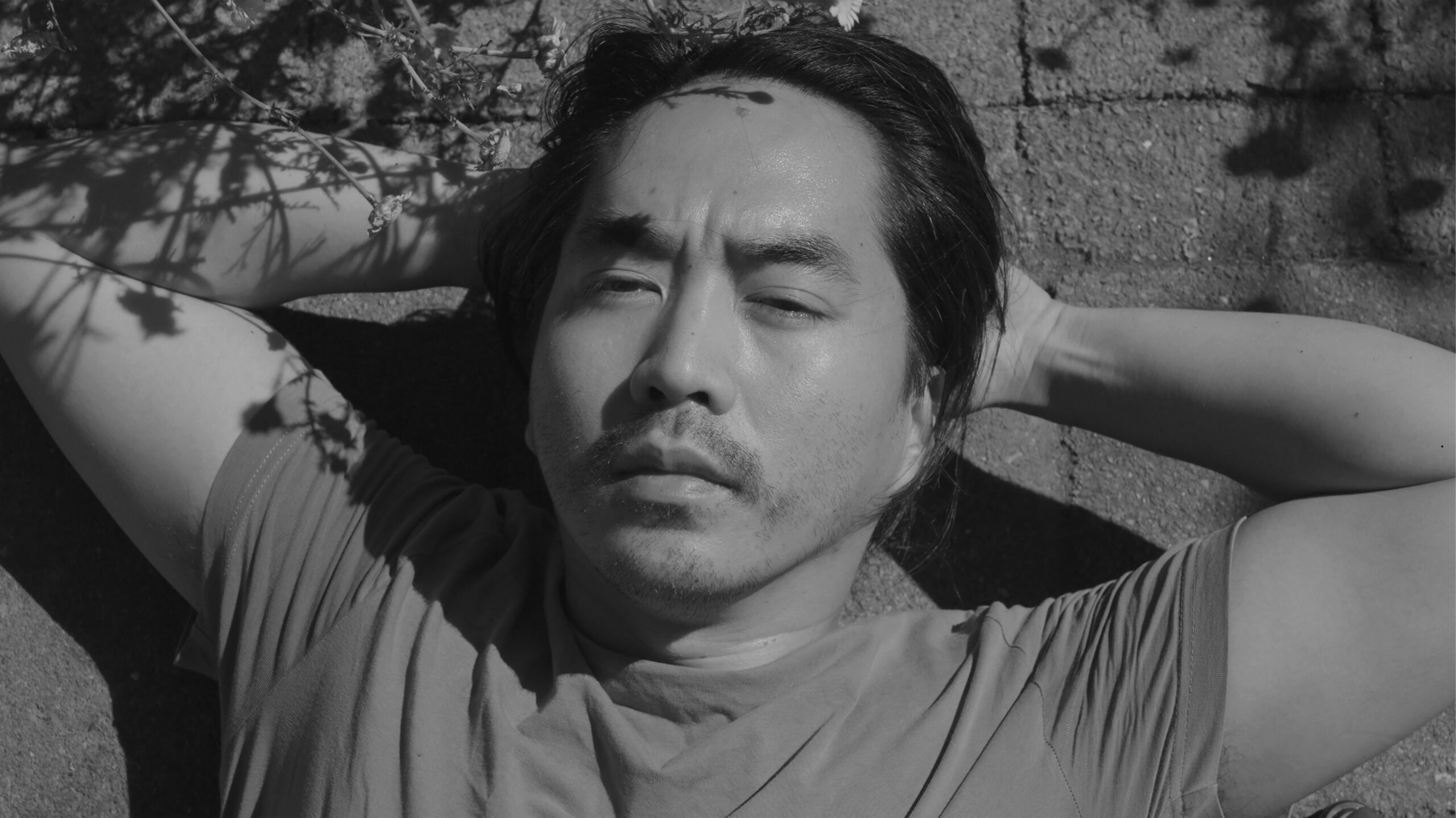Close up of a young man with his hands behind his head, shadows of foliage on his arm, gazing up at the sky