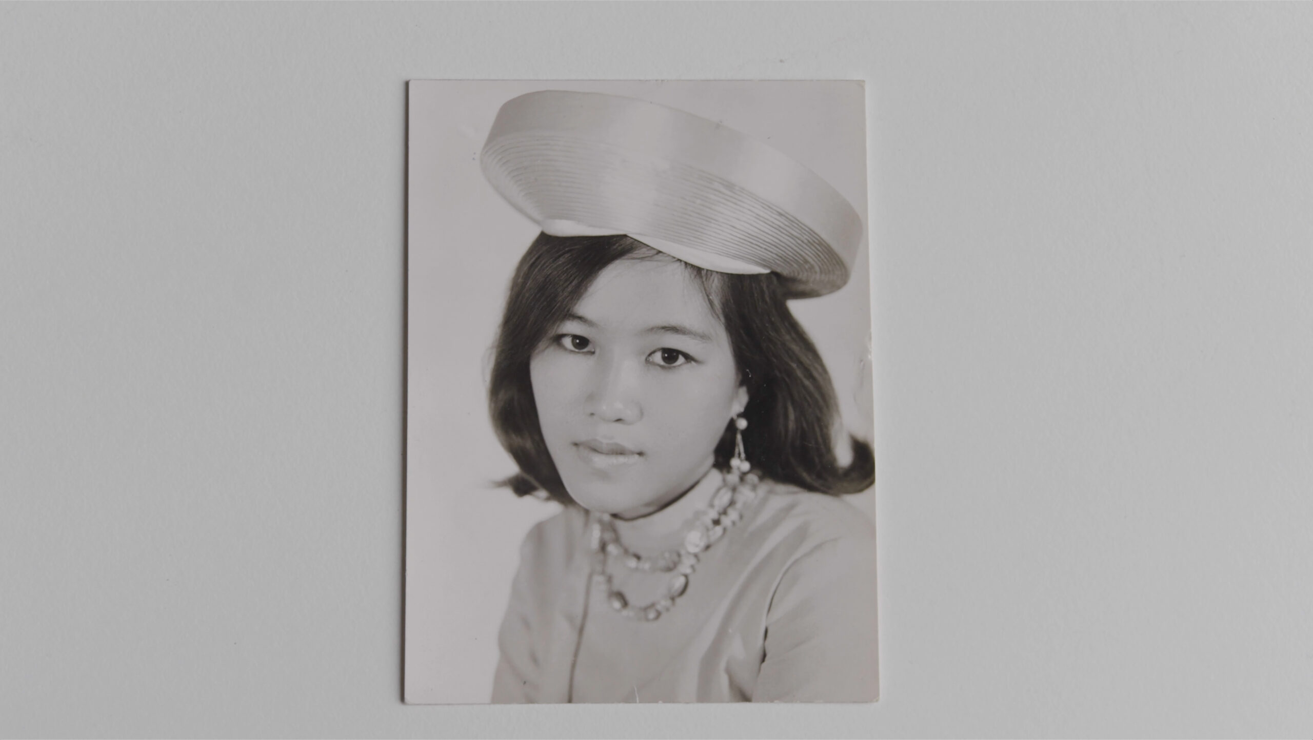 Black-and-white sixties era photograph of a young woman in a circular silk hat, beaded necklace and earrings, and mock-neck top posing for a portrait
