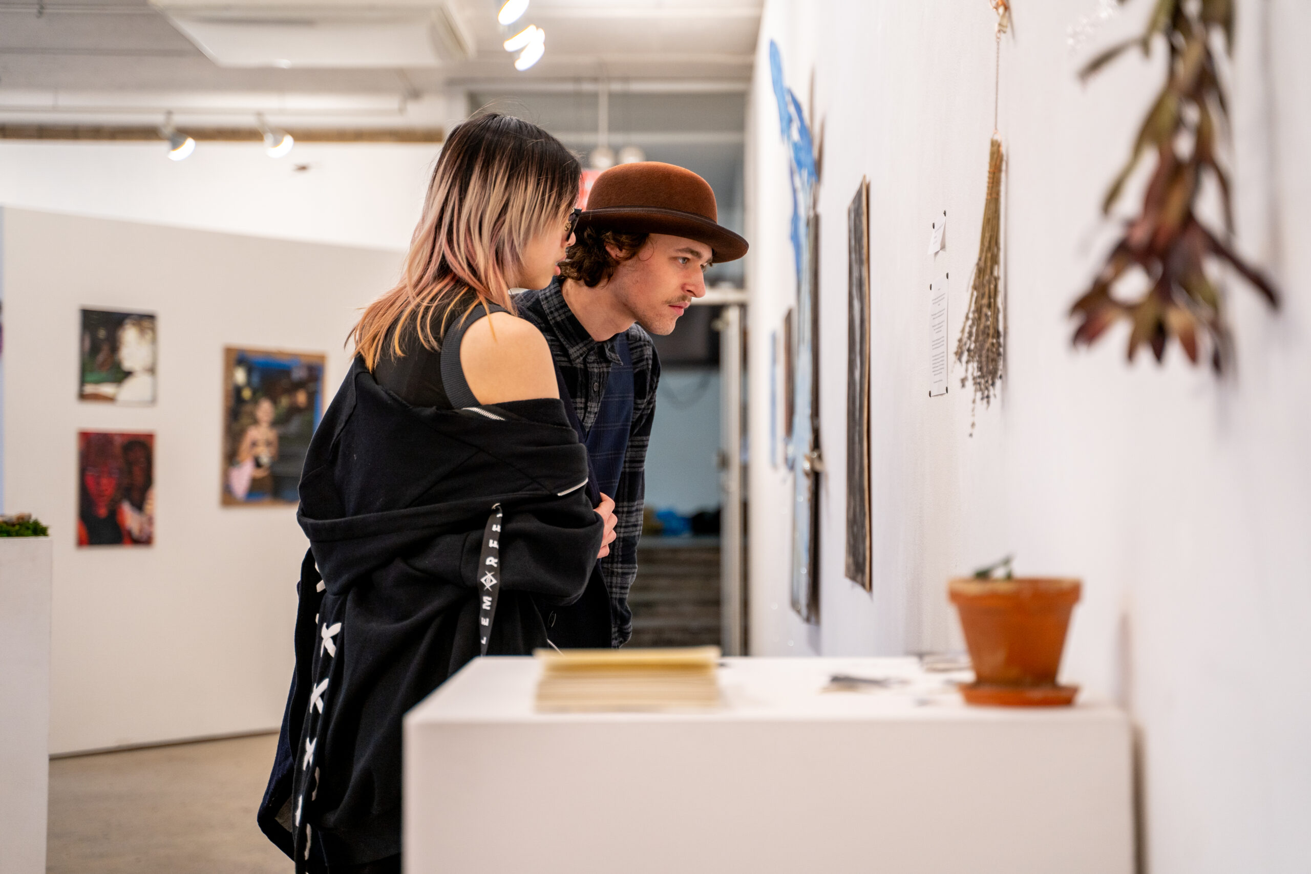 Two gallery visitors lean in to view wall-mounted artworks and hanging dried plants in a well-lit exhibition space, with additional framed art visible in the background.