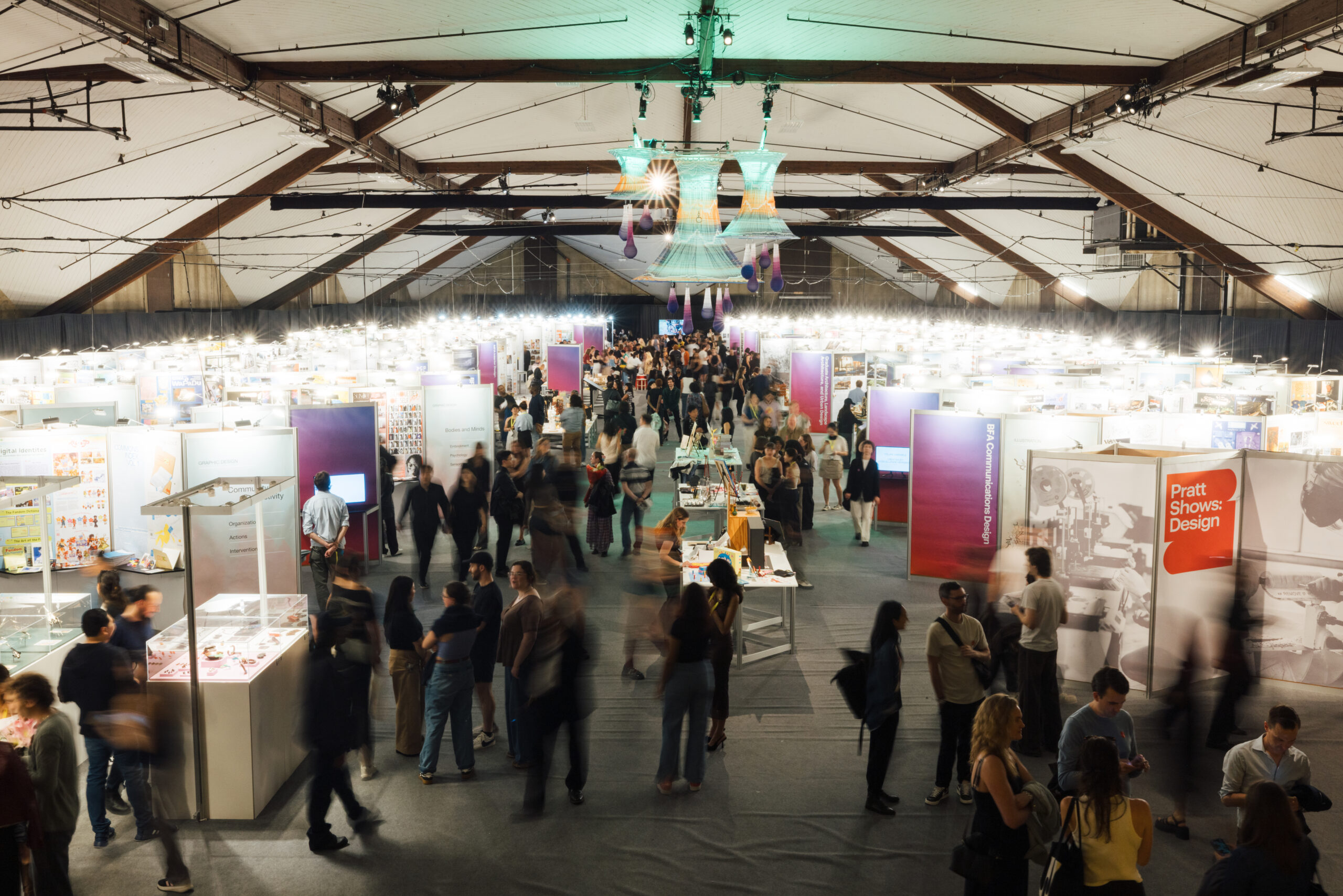 Wide overhead view of a crowded indoor design exhibition with rows of display booths and signage, as visitors walk through aisles beneath a high arched ceiling and hanging installation.