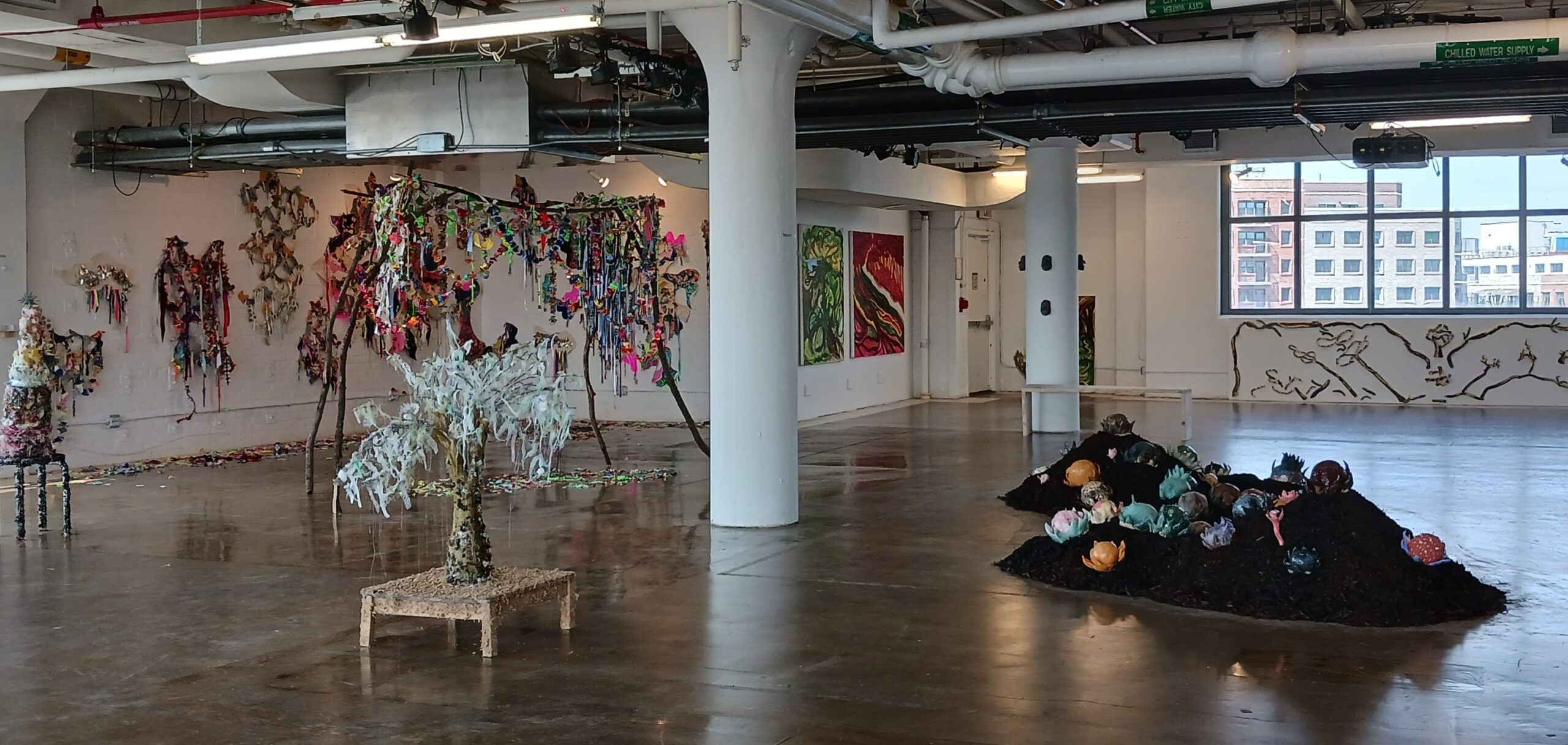 view of open gallery room, large pillar in the middle of room, with sculptures installed in the foreground, drawings and paintings on wall in the background