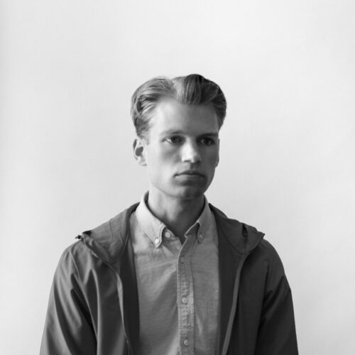 Josh Skirtich stands in front of a blank background, wearing a casual jacket and button-down shirt, looking serious.