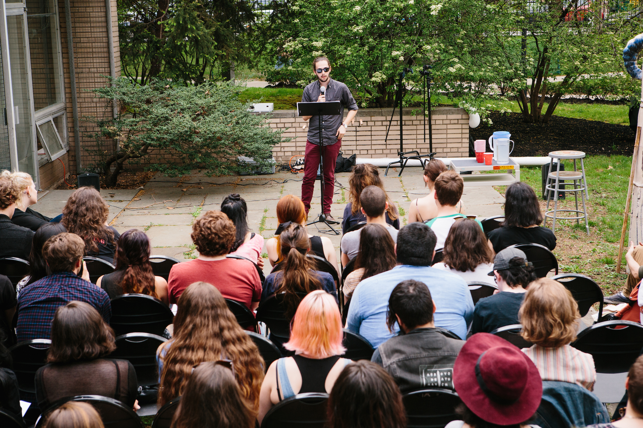 student speaking to group of about twenty, on campus grounds, standing at podium