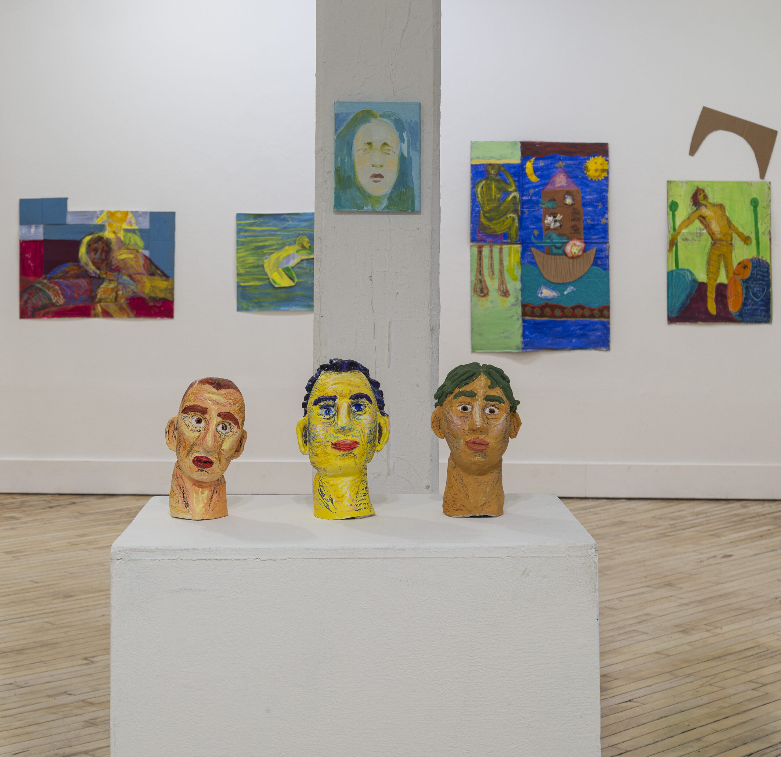 sculptures of faces on a display table in foreground, gallery room with paintings similar to the sculptures in background