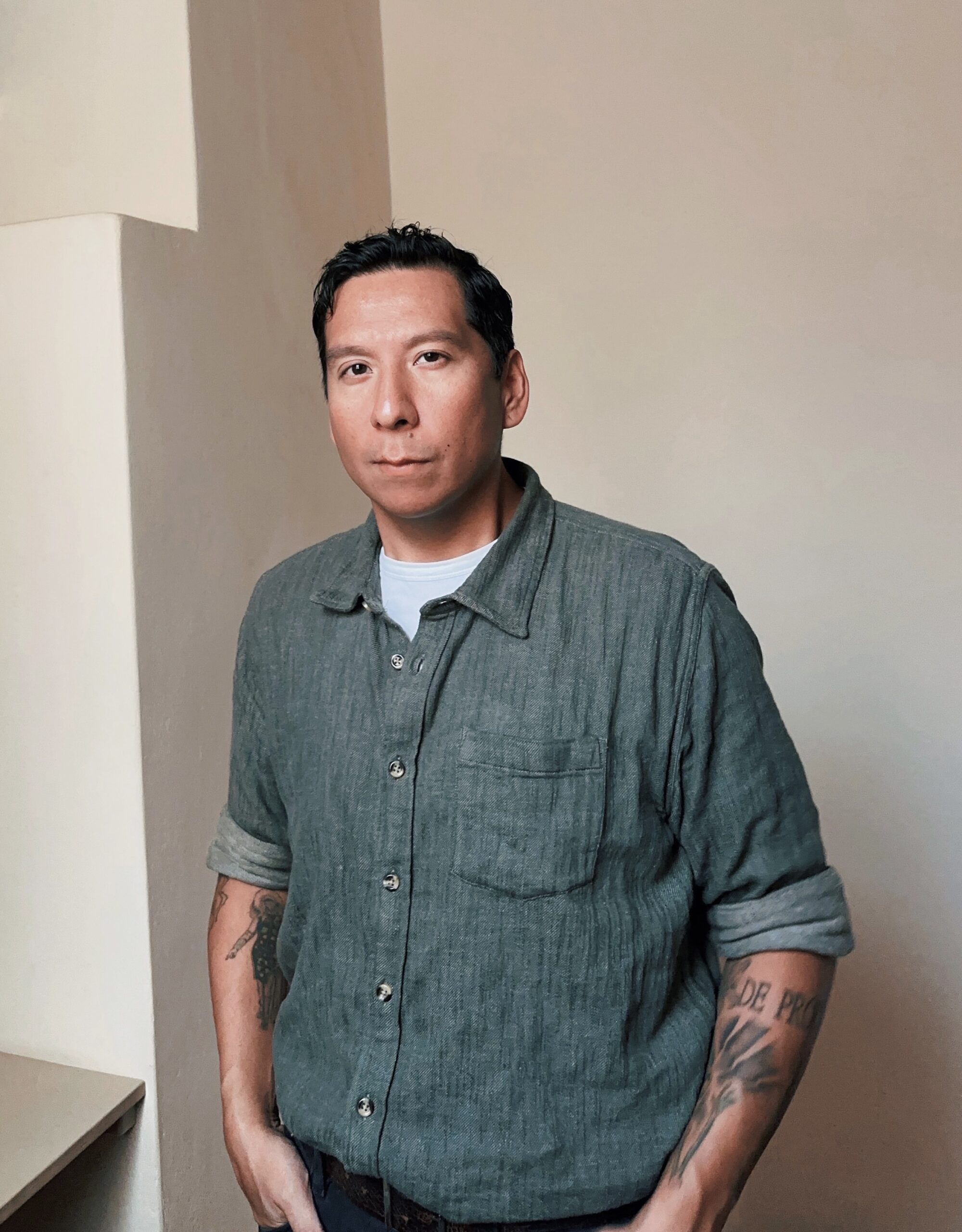 An image of Sky Hopinka standing in front of a beige wall in a teal button-down shirt.
