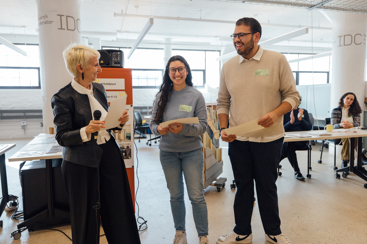 two students, smiling, standing with president Bronet, holding award documentation, with offices in background