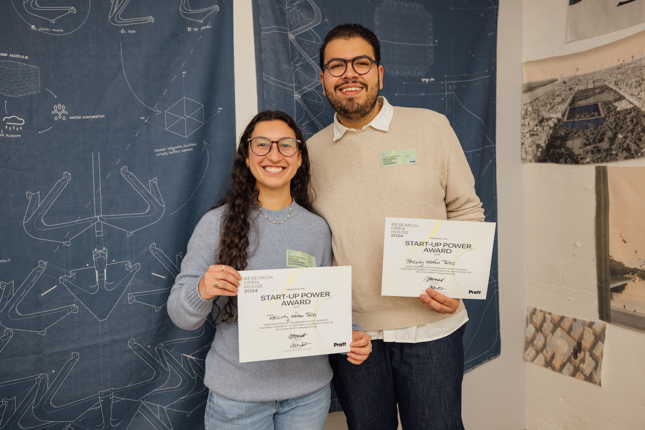 two students, smiling, holding award documentation, with tapestry in background