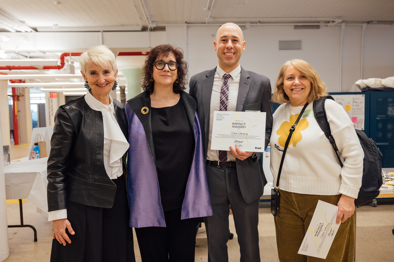 three faculty members, smiling, holding award documentation, standing with president Bronet, with offices in background