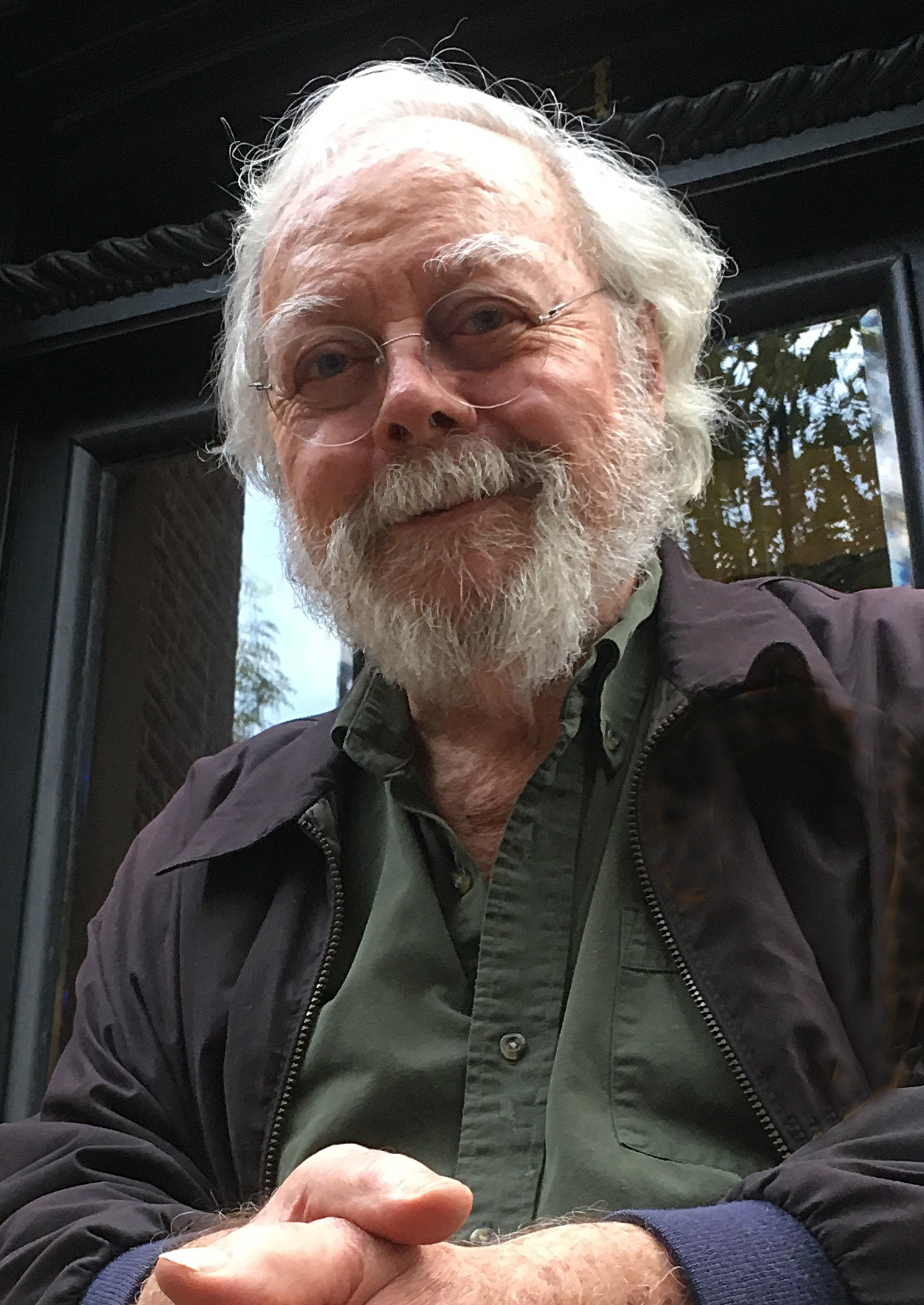 Man with white hair and beard smiles, posed with hands clasped