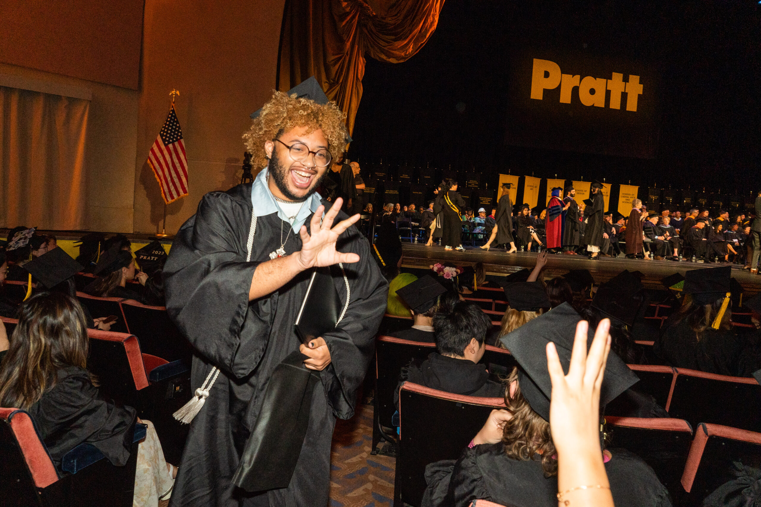 student waving at another student, wearing commmencement cap and gown, inside of radio city music hall