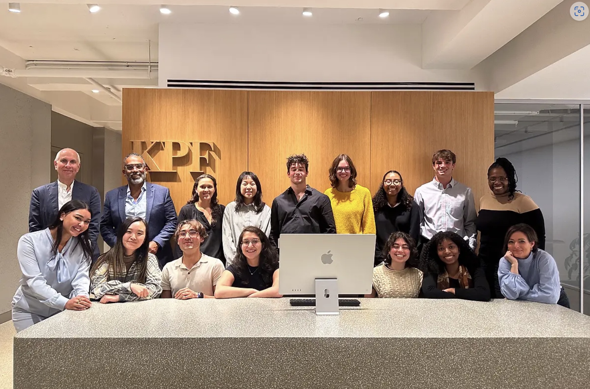 A group photo of people standing behind a table, with the letters KPF on the wall behind them.