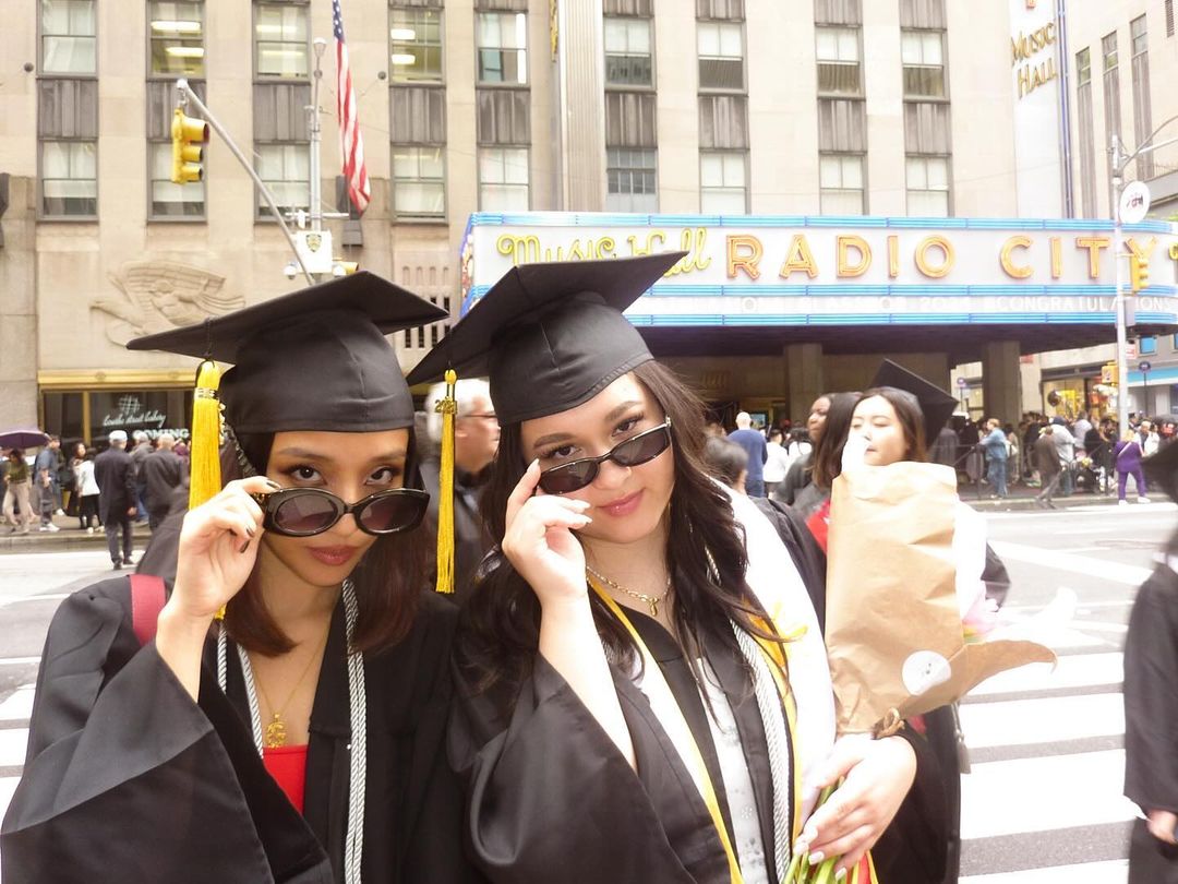Two people in their commencement cap and gowns holding their sunglasses on their noses posing in front of the exterior of Radio City Music Hall. Person on the right is holding pink flowers.
