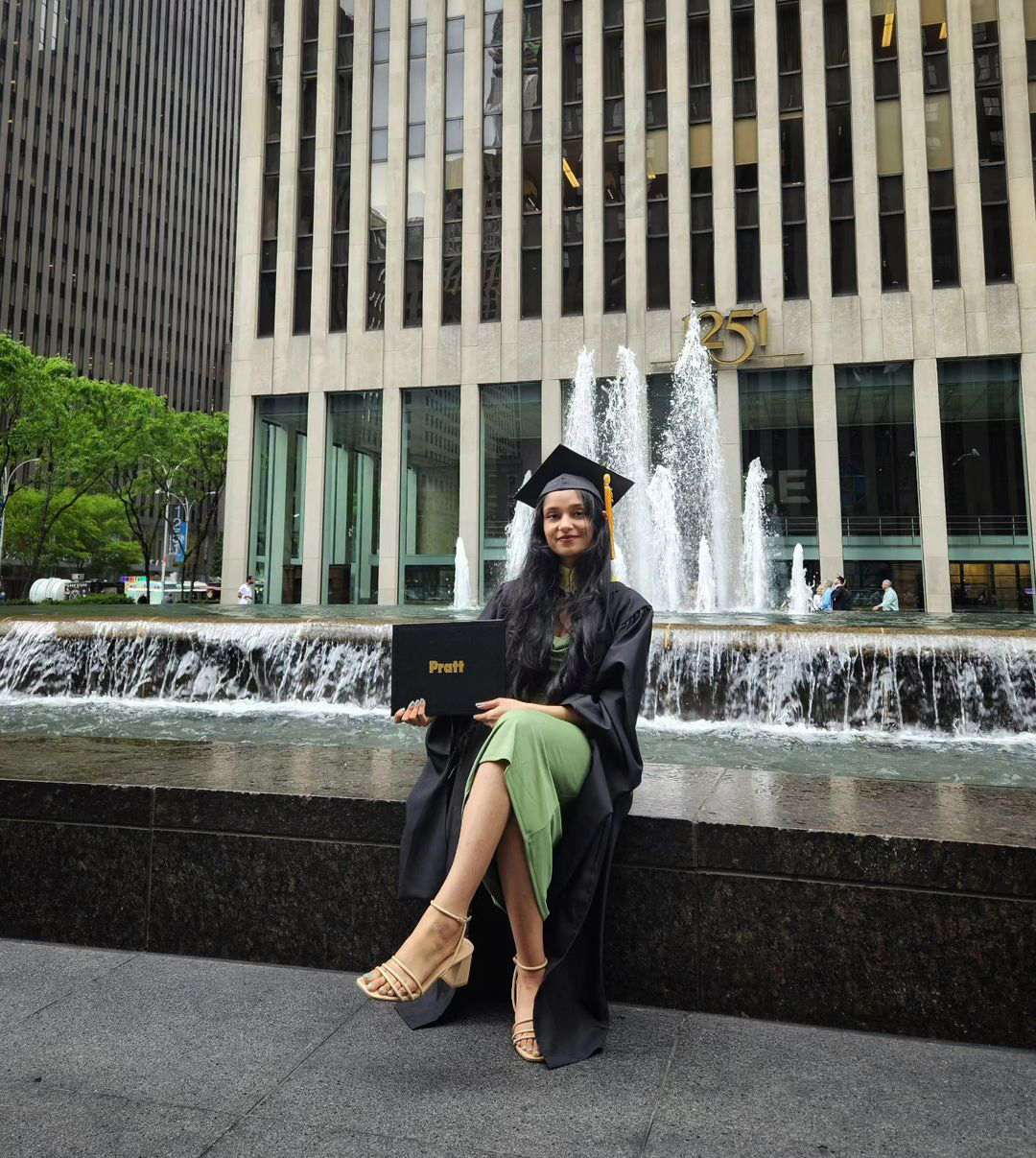 Person in a green dress and commencement cap and gown sitting in front of a fountain, smiling and holding up their Pratt diploma.