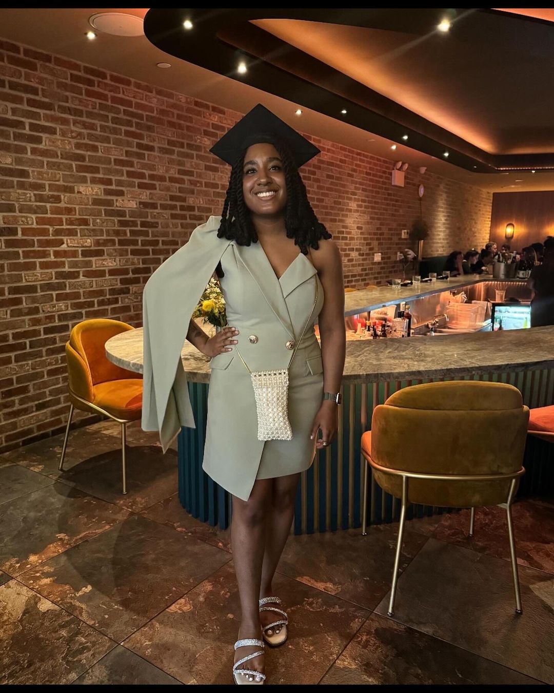 Person standing in a beige dress and commencement cap smiling inside of a restaurant.