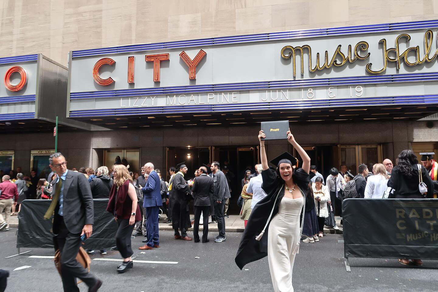 Person in a white dress and commencement cap and gown smiling and holding up their Pratt diploma in front of the exterior of Radio City Music Hall.